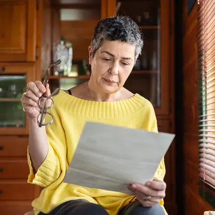 Mature woman holding her glasses and reading a letter, sitting near window with blinds in her home