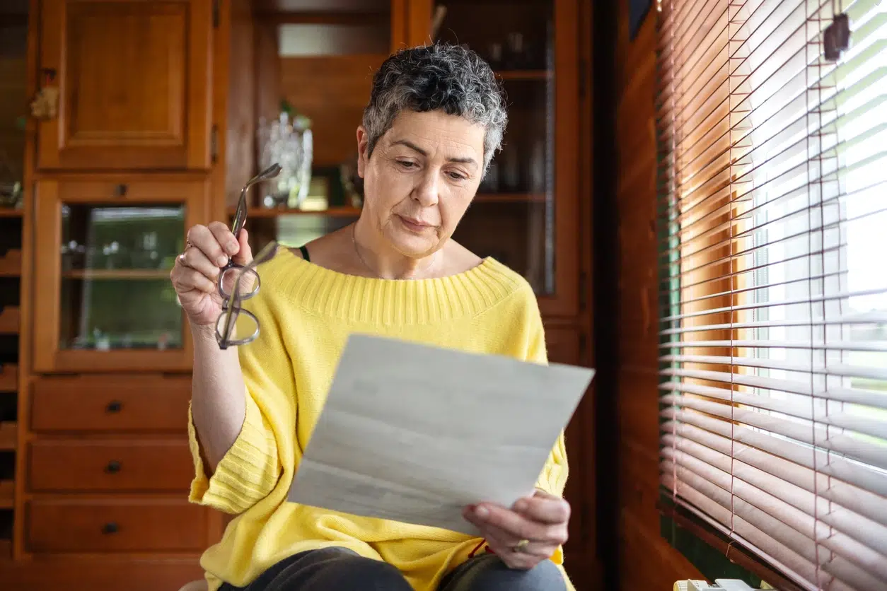 Mature woman holding her glasses and reading a letter, sitting near window with blinds in her home